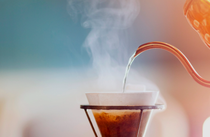 Fotografia em close de água quente sendo despejada por uma chaleira de bico fino sobre um coador tipo V60 com filtro de papel, com vapor subindo e café sendo extraído ao fundo, em fundo desfocado azul e laranja suave.