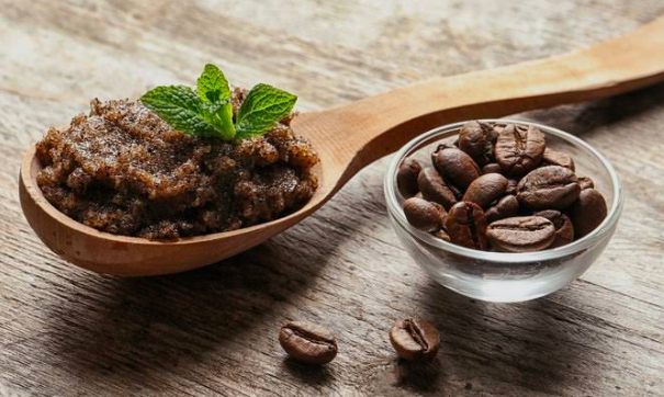 Colher de madeira com esfoliante de borra de café decorada com folhas de hortelã fresca, ao lado de uma tigela de vidro com grãos de café torrado, sobre superfície de madeira rústica.