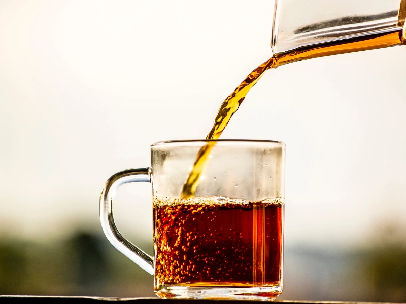 Fotografia em close de café líquido sendo despejado de uma jarra em uma xícara de vidro transparente, com a bebida em tom âmbar avermelhado e bolhas se formando na superfície, em fundo claro desfocado.