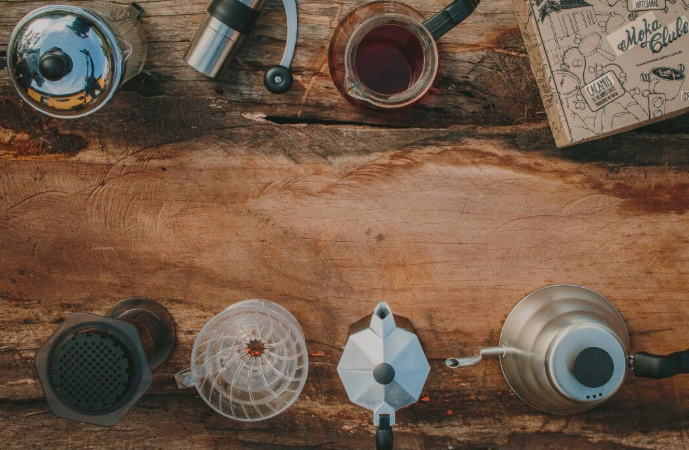 Vista de cima de uma mesa de madeira rústica com diversos equipamentos de preparo de café dispostos ao redor: coador tipo Hario, cafeteira italiana (moka), chaleira, moedor manual, prensa francesa, xícara com café e uma caixa decorativa, ilustrando diferentes métodos de preparo.