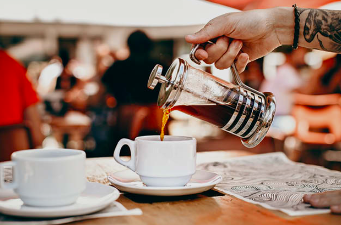Mão com tatuagem despejando café de uma prensa francesa metálica em uma xícara branca sobre pires, em ambiente de cafeteria ao ar livre com pessoas e cadeiras coloridas desfocadas ao fundo.

