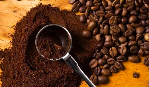 Superfície de madeira com montinho de café moído fino, uma colher dosadora de metal mergulhada no pó, e grãos de café torrado espalhados ao lado, em composição que ilustra o processo de moagem do café.