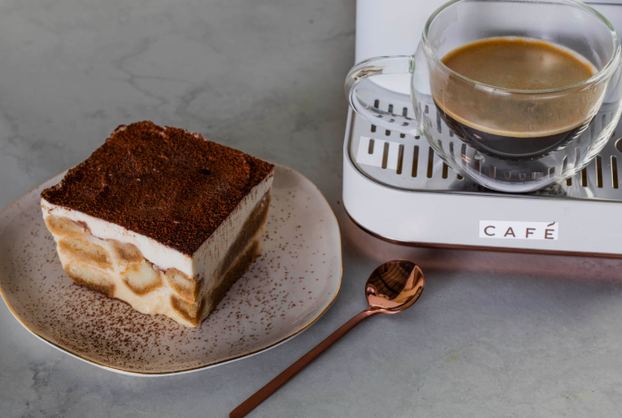 Fatia de tiramisù com camadas visíveis de biscoito embebido em café e creme, coberta com cacau em pó, servida em prato de cerâmica ao lado de uma xícara de vidro com espresso e uma colher de cobre, sobre bancada cinza.