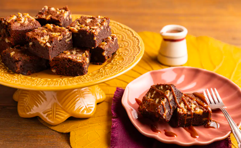 Pedaços de brownie de chocolate e café dispostos em um prato de bolo amarelo, com uma fatia servida em prato rosa ao lado com garfo, sobre mesa de madeira com guardanapo colorido e potinho de calda ao fundo.