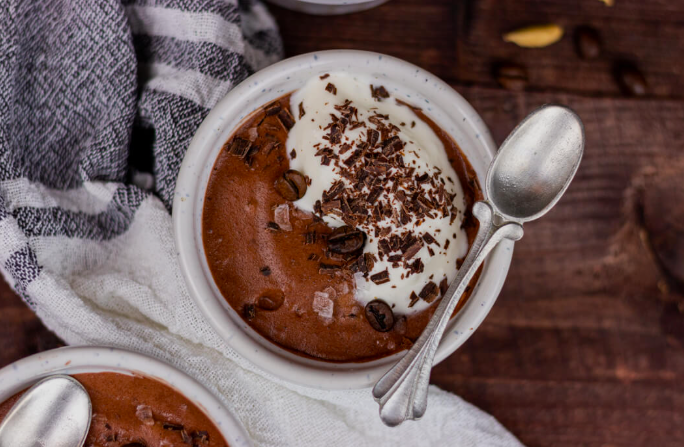Tigela branca de cerâmica com mousse de chocolate escuro, coberta com chantilly, raspas de chocolate e grãos de café, acompanhada de duas colheres de prata, sobre mesa de madeira rústica com pano xadrez ao fundo.