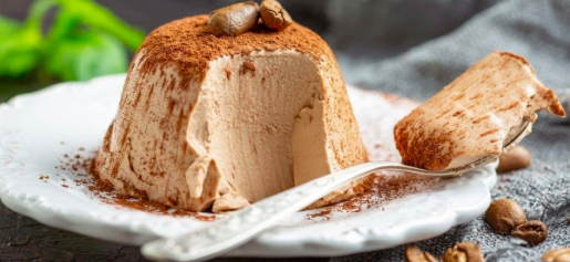 Panna Cotta de café em formato de cúpula servido em prato branco com bordas rendadas, coberto com cacau em pó e decorado com grãos de café, com uma fatia cortada ao lado e uma colher, sobre fundo acinzentado.