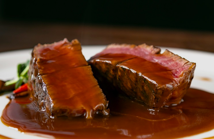Duas fatias de filé mignon ao ponto, com interior rosado e crosta escura, servidas em prato branco com molho escuro e brilhante à base de café, acompanhadas de legumes ao fundo, em apresentação de restaurante com iluminação dramática.